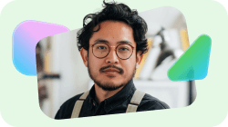 A man with dark curly hair, a beard, and round glasses, wearing a dark shirt and suspenders, looks forward.