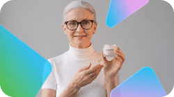 A smiling woman with grey hair and glasses holds up a jar of cosmetic cream.