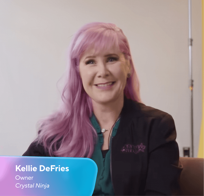 Kellie DeFries, owner of Crystal Ninja, with pink hair, smiling and wearing a black jacket with a purple Crystal Ninja logo.