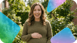 A smiling pregnant woman in a green jumper holds her belly, surrounded by trees.